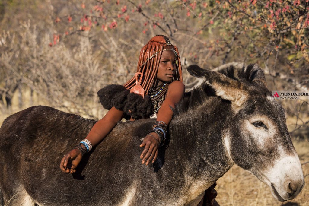 Angola - Una donna Himba con il suo asino. Le donne himba sono famose perché coprono il loro corpo con l'otjize, una mistura cosmetica fatta di burro e ocra. Questo impasto assicura la pulizia della pelle per un lungo tempo, e protegge la pelle dal clima estremamente arido e secco del Kaokoland, oltre che dalle punture delle zanzare e di altri insetti